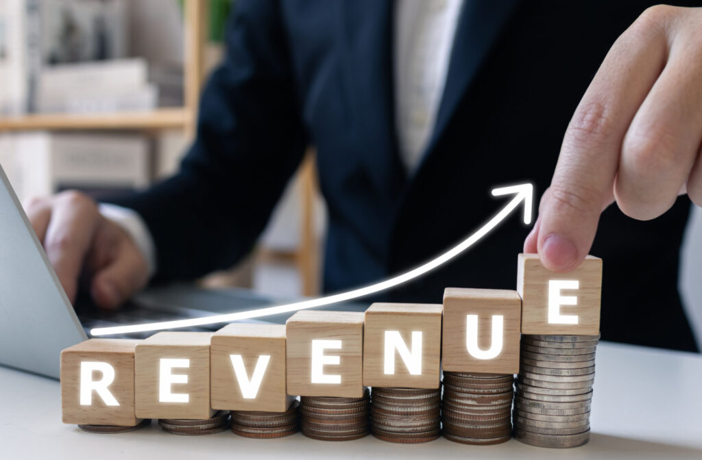 A stack of coins on a table that grows from small to large with blocks on top that spell out "REVENUE" in glowing letters. A man in a suit is placing the final letter with his left hand.