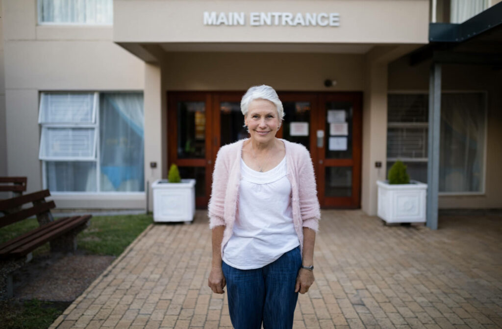 A smiling older woman standing in front of the entrance to a senior living community