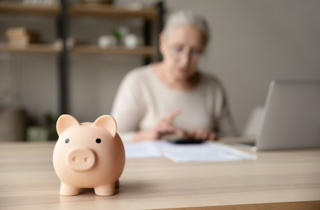 Close-up of a pink piggy bank sitting on a table with an older woman out of focus in the background, using a laptop and a calculator.