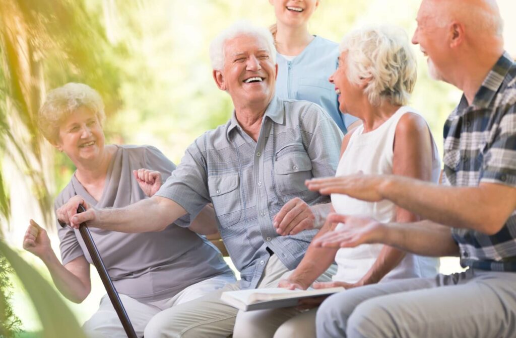 An older adult man with a cane smiles and laughs while sitting outside with a nurse and other seniors.