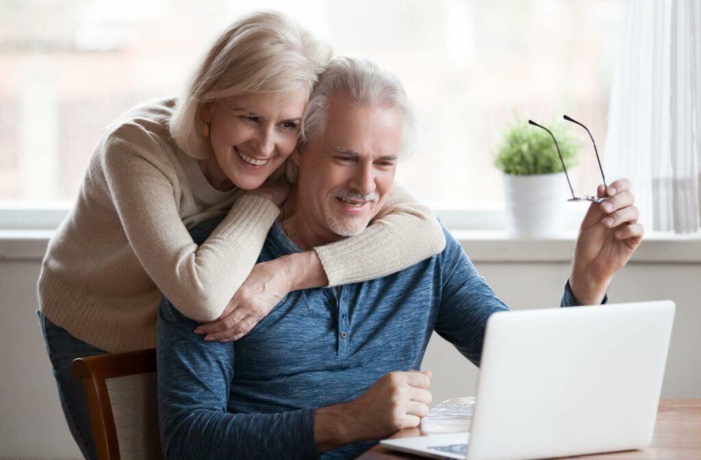 Older adults smiling while researching senior living options on a laptop, sitting on a living room sofa in a bright setting