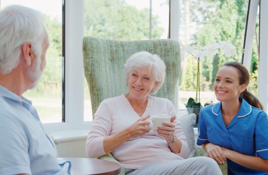 A senior holding a mug smiles to their loved one on their left while a caregiver crouches next to them in a bright skilled nursing home lounge